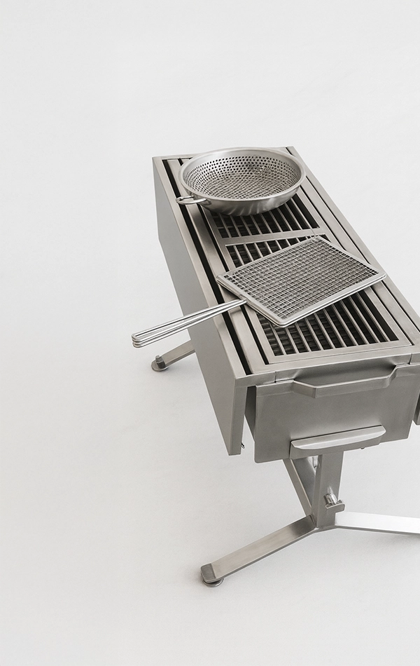 Barbacoa alta de acero inoxidable con parrilla central y menaje de malla colocado encima vista en formato vertical.