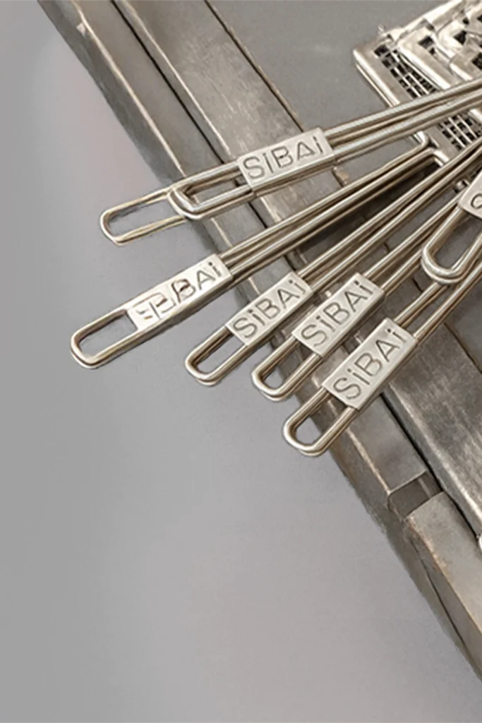 Detalle de varias varillas o asas de acero inoxidable con el logo SiBAi grabado, apoyadas sobre la estructura metálica de una parrilla.